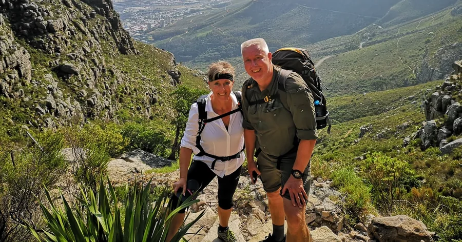 A smiling couple with daypacks on a rocky trail, aloes beside them and the city far below