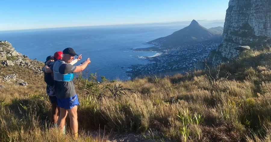 Two hikers on a grassy slope photographing Lion's Head and the Atlantic Ocean
